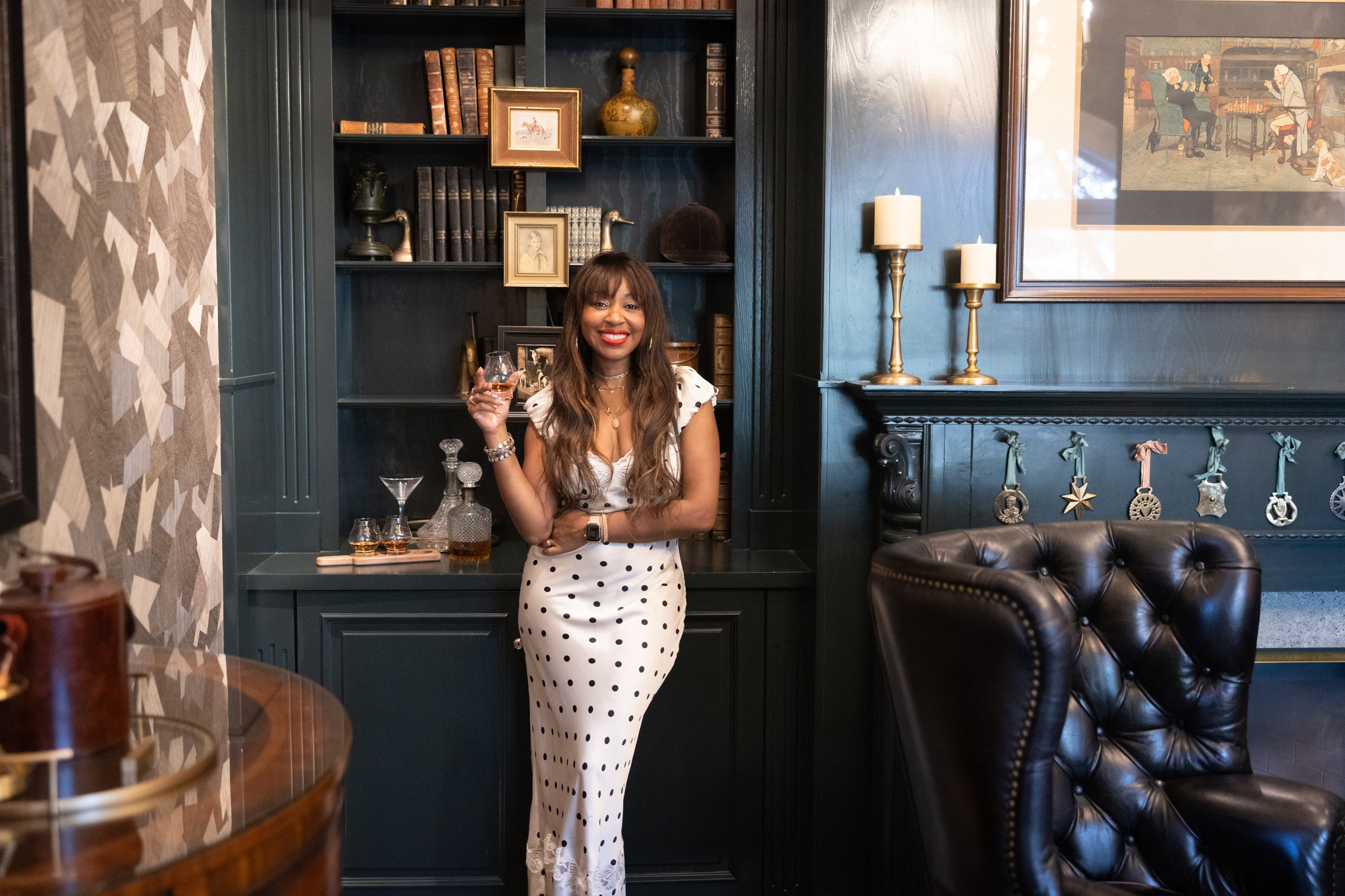 Woman in polka dot dress holding a drink in a sophisticated study with bookshelves, candles, and leather armchair.