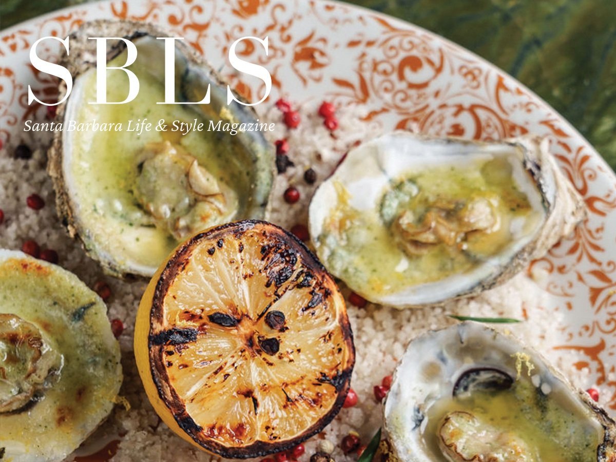 Grilled oysters with herb butter and half a grilled lemon on a decorative plate, featured in Santa Barbara Life & Style Magazine.