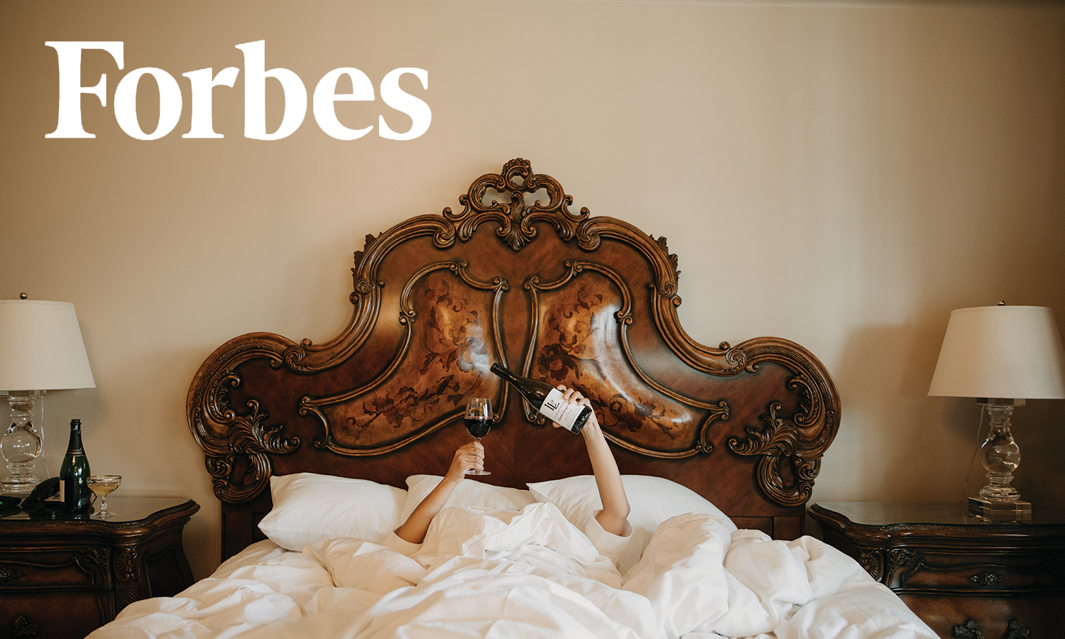 Person in bed under a comforter holding a wine bottle and glass, ornate headboard, Forbes logo, wine glass on side table.